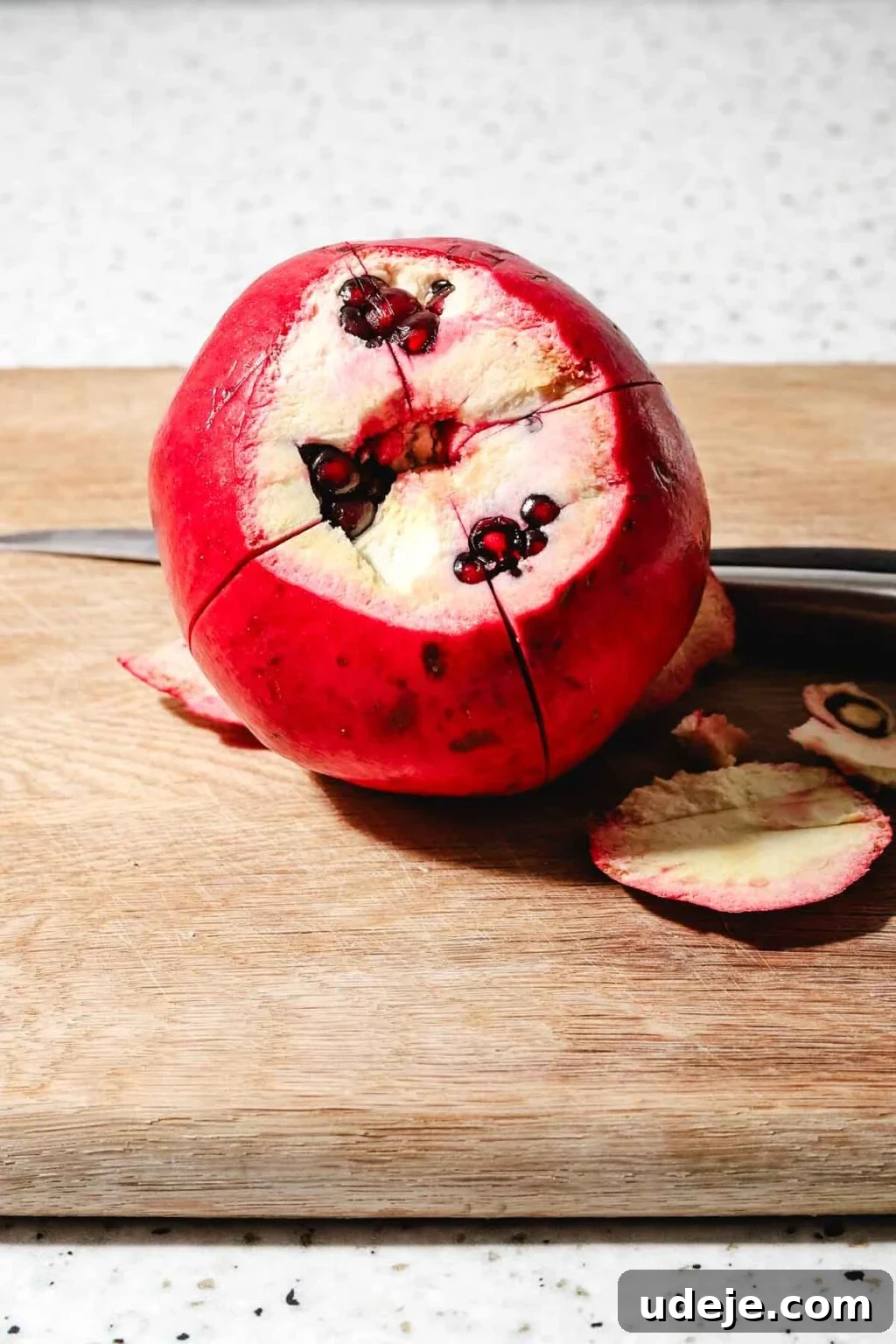 Homemade Pomegranate Molasses: Sweet, Tangy, and Versatile 9 Pomegranate with the top sliced off and some pomegranate seeds showing, with four cuts crossing each other going through it on a wooden cutting board with a knife beside it on a speckled background