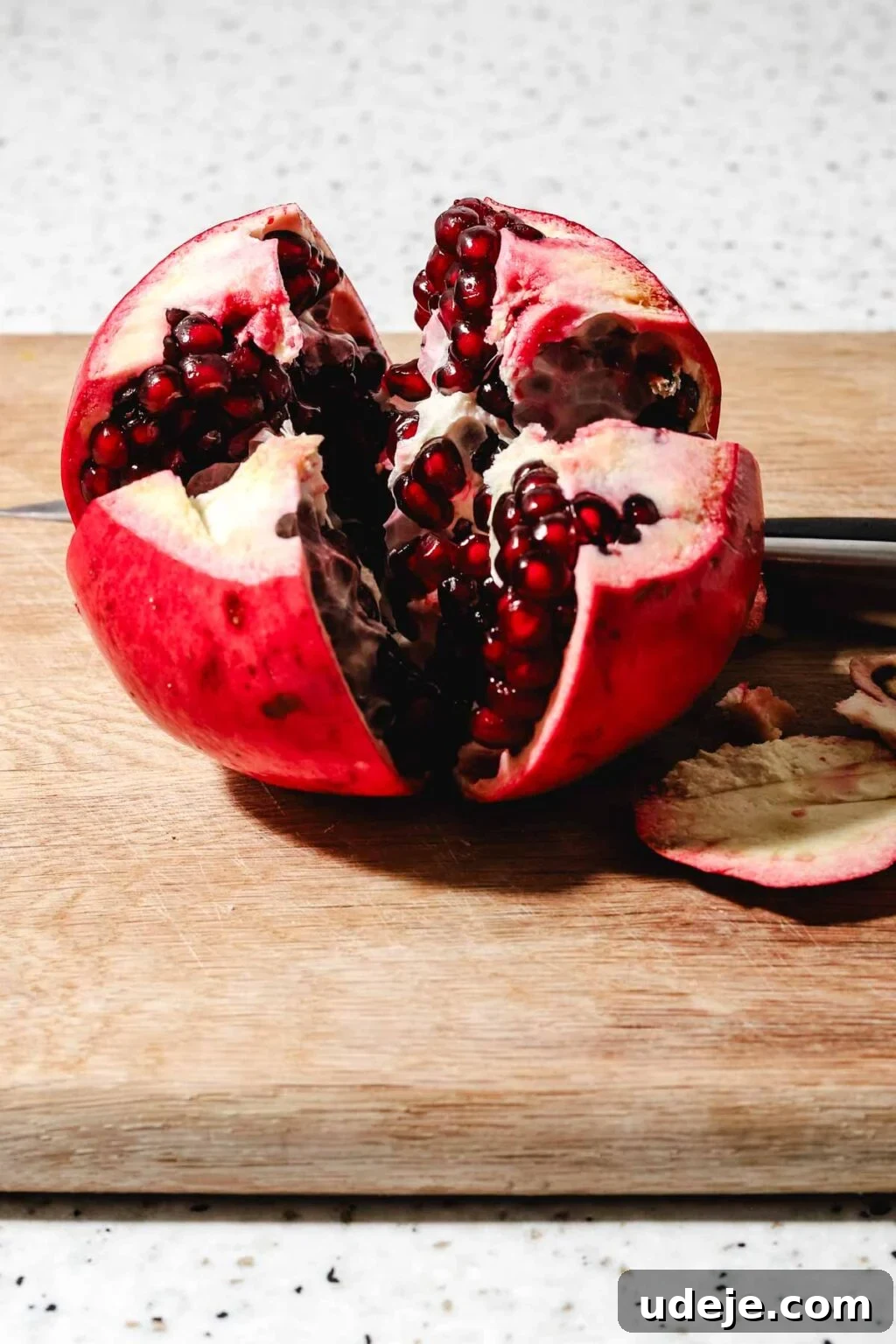 Homemade Pomegranate Molasses: Sweet, Tangy, and Versatile 10 Pomegranate cut into four sections but still attached at the bottom on a wooden cutting board with a knife at the back on a speckled background