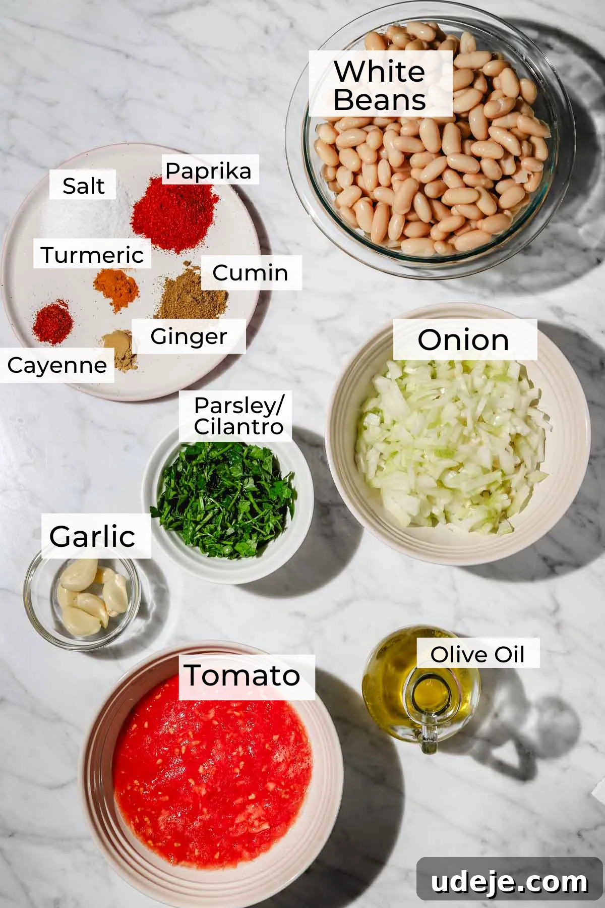 Moroccan Loubia A Flavorful Vegetarian White Bean Stew 3 Ingredient shot for Moroccan Loubia with white beans in a glass bowl, paprika, salt, turmeric, cumin, ginger and cayenne on a small white plate, chopped onions in a white bowl, parsley/cilantro in a small white bowl, garlic cloves in a small glass bowl, olive oil in a small glass pitcher and tomato in a white bowl