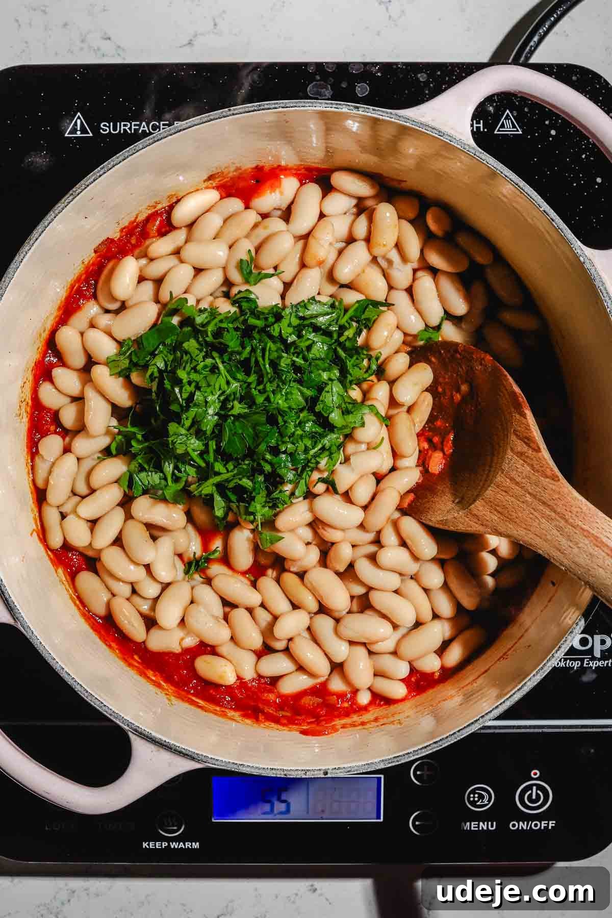 Moroccan Loubia A Flavorful Vegetarian White Bean Stew 7 Sautéed chopped onions, tomatoes and spices with white beans on top and chopped parsley n a large beige pot with a wooden spoon on a marble