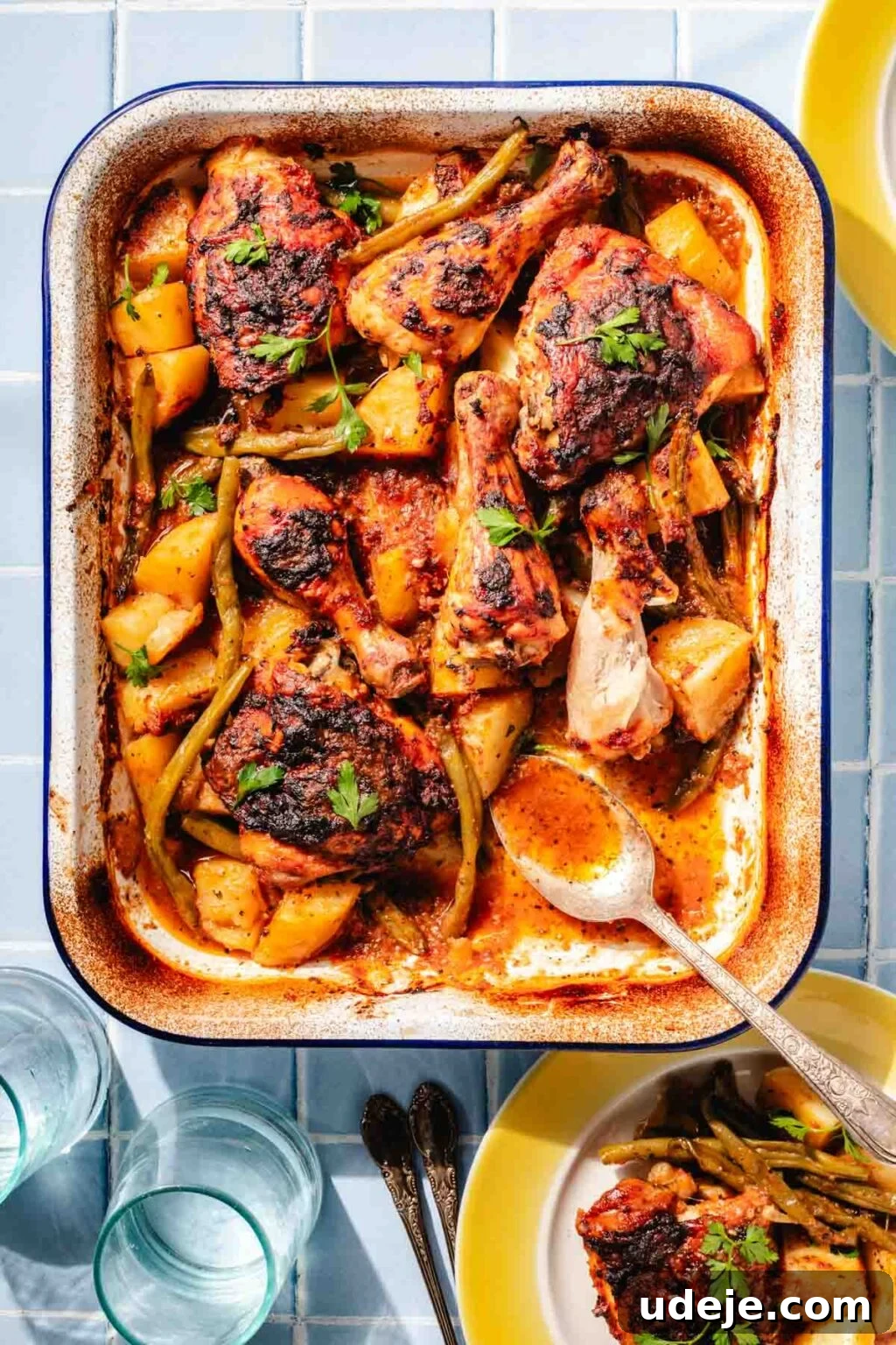 Irresistible Mediterranean Oven-Roasted Chicken 2 Baked Mediterranean chicken with potatoes and green beans in a white and blue rimmed baking dish with a metal spoon, a yellow rimmed plate with a serving of Mediterranean chicken, and 2 water glasses on a blue tiled background