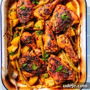 Baked mediterranean chicken with potatoes and green beans in a white and blue rimmed baking dish with a metal spoon on a blue tiled background