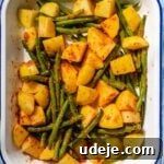 cubed potatoes and trimmed green beans mixed with blended spice mixture in a white and blue rimmed baking pan on a blue tiled background