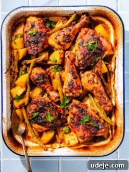 Baked mediterranean chicken with potatoes and green beans in a white and blue rimmed baking dish with a metal spoon on a blue tiled background