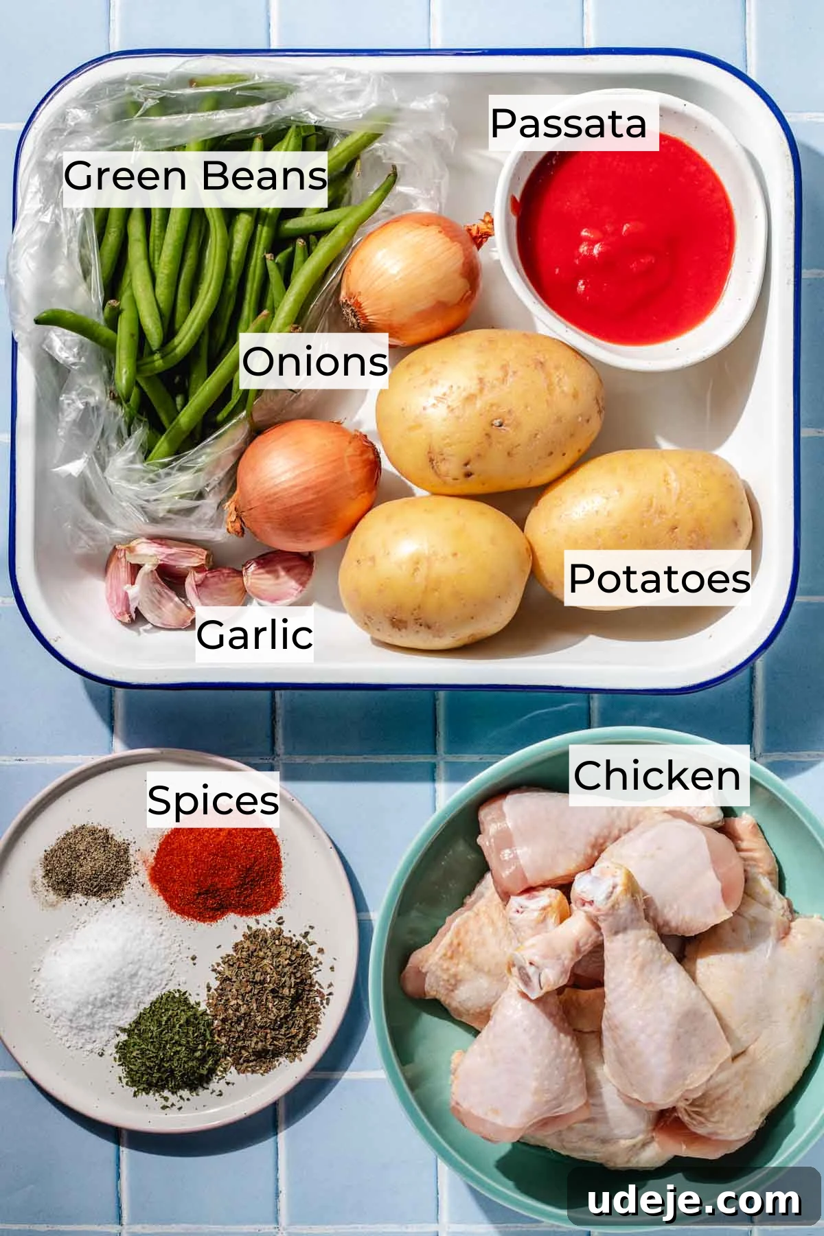 Irresistible Mediterranean Oven-Roasted Chicken 3 Ingredient shot for Mediterranean chicken with a white blue rimmed baking dish with passata in a white bowl, green beans in a bag, onions, potatoes and garlic, a small plate with spices and a mint coloured bowl with chicken legs on a blue tiled background