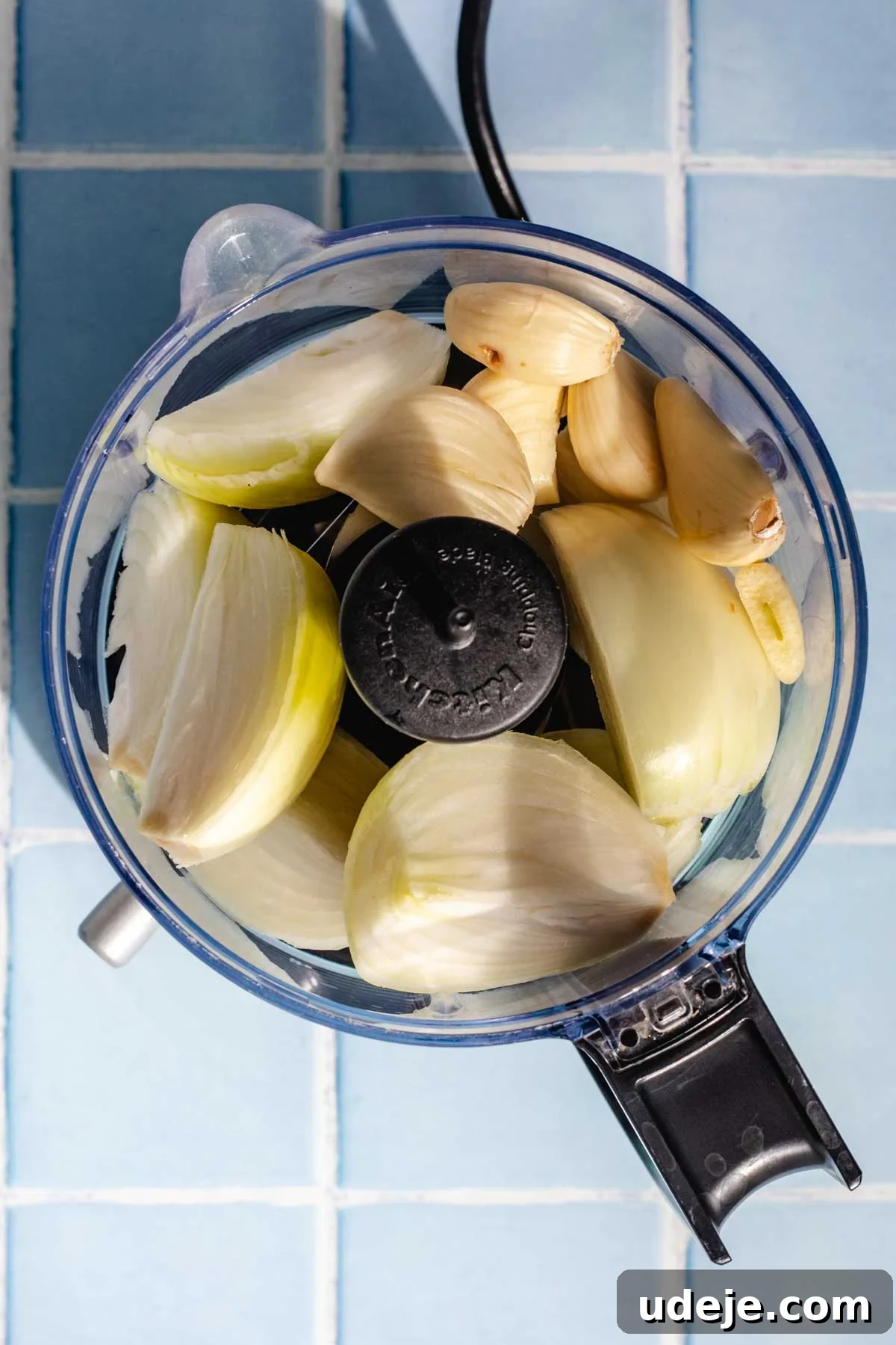 Irresistible Mediterranean Oven-Roasted Chicken 4 Quartered and peeled white onions and peeled garlic cloves in a food processor on a blue tiled background