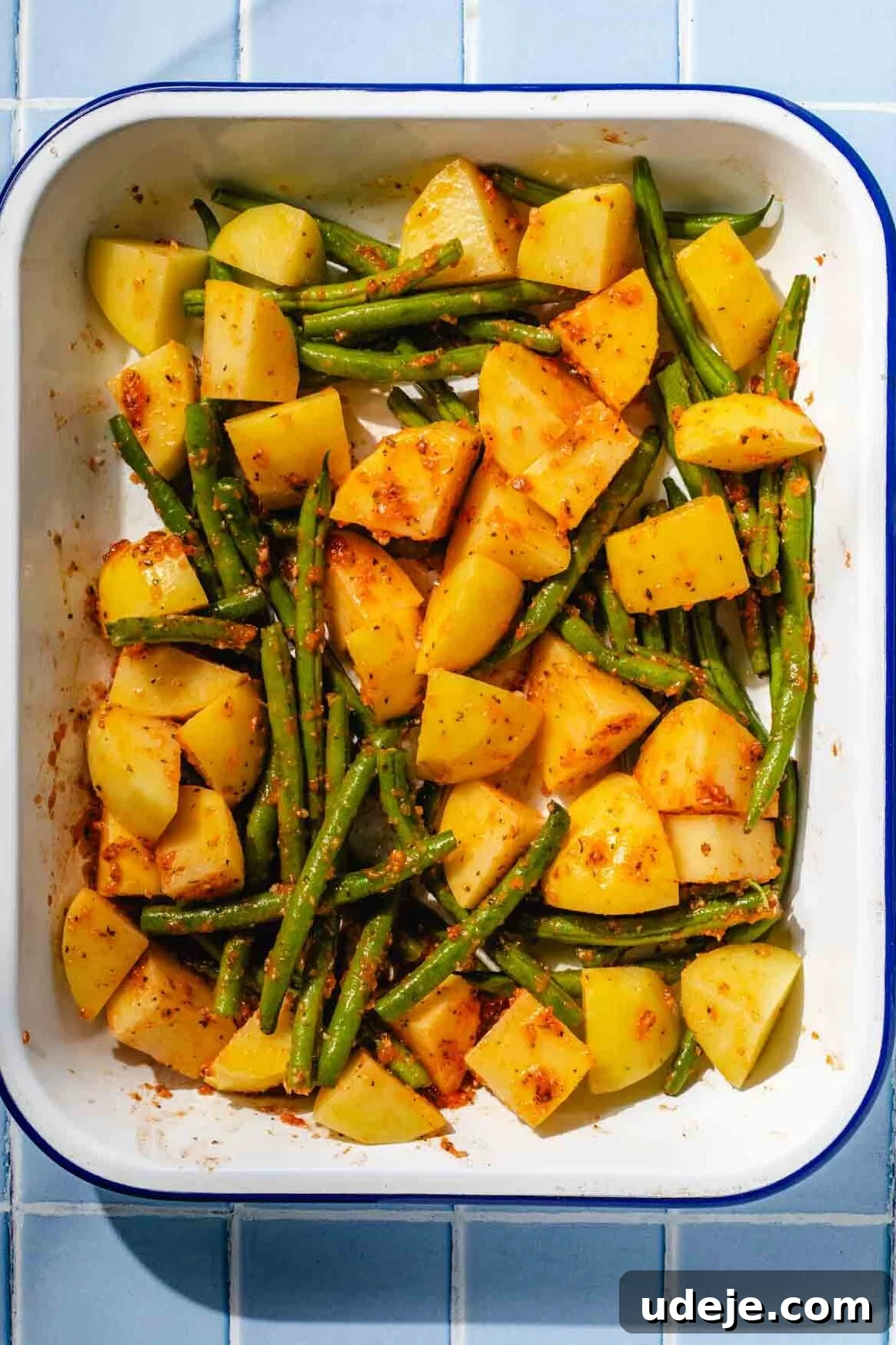 Irresistible Mediterranean Oven-Roasted Chicken 7 cubed potatoes and trimmed green beans mixed with blended spice mixture in a white and blue rimmed baking pan on a blue tiled background