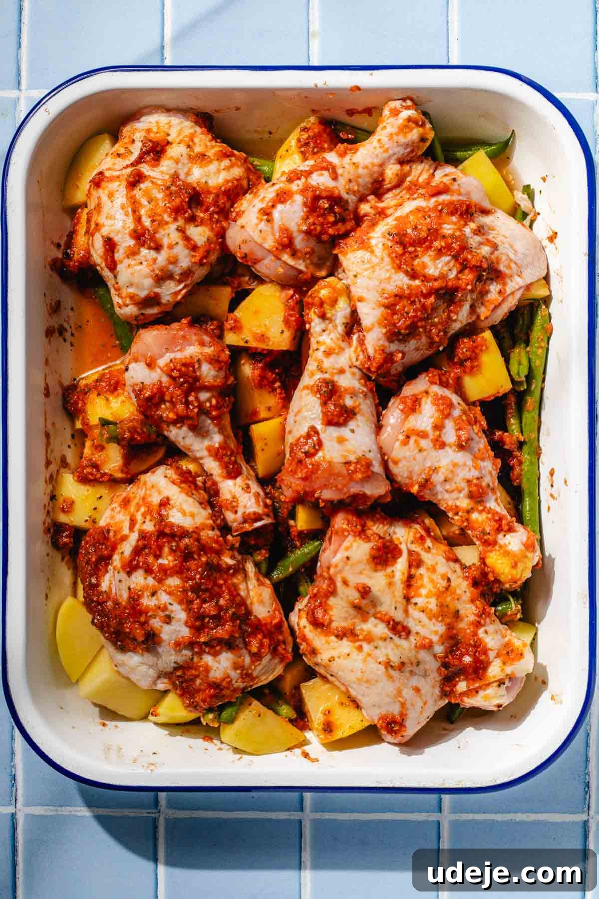 Irresistible Mediterranean Oven-Roasted Chicken 8 Chicken, cubed potatoes and trimmed green beans mixed with blended spice mixture in a white and blue rimmed baking pan on a blue tiled background
