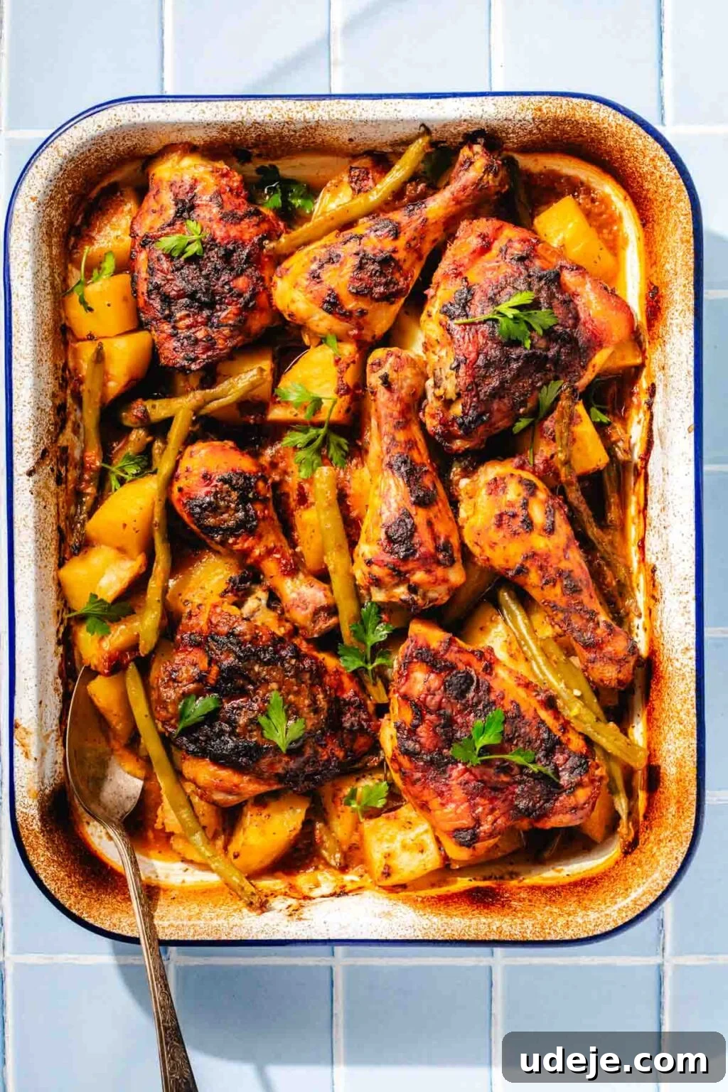 Irresistible Mediterranean Oven-Roasted Chicken 9 Baked Mediterranean chicken with potatoes and green beans in a white and blue rimmed baking dish with a metal spoon on a blue tiled background