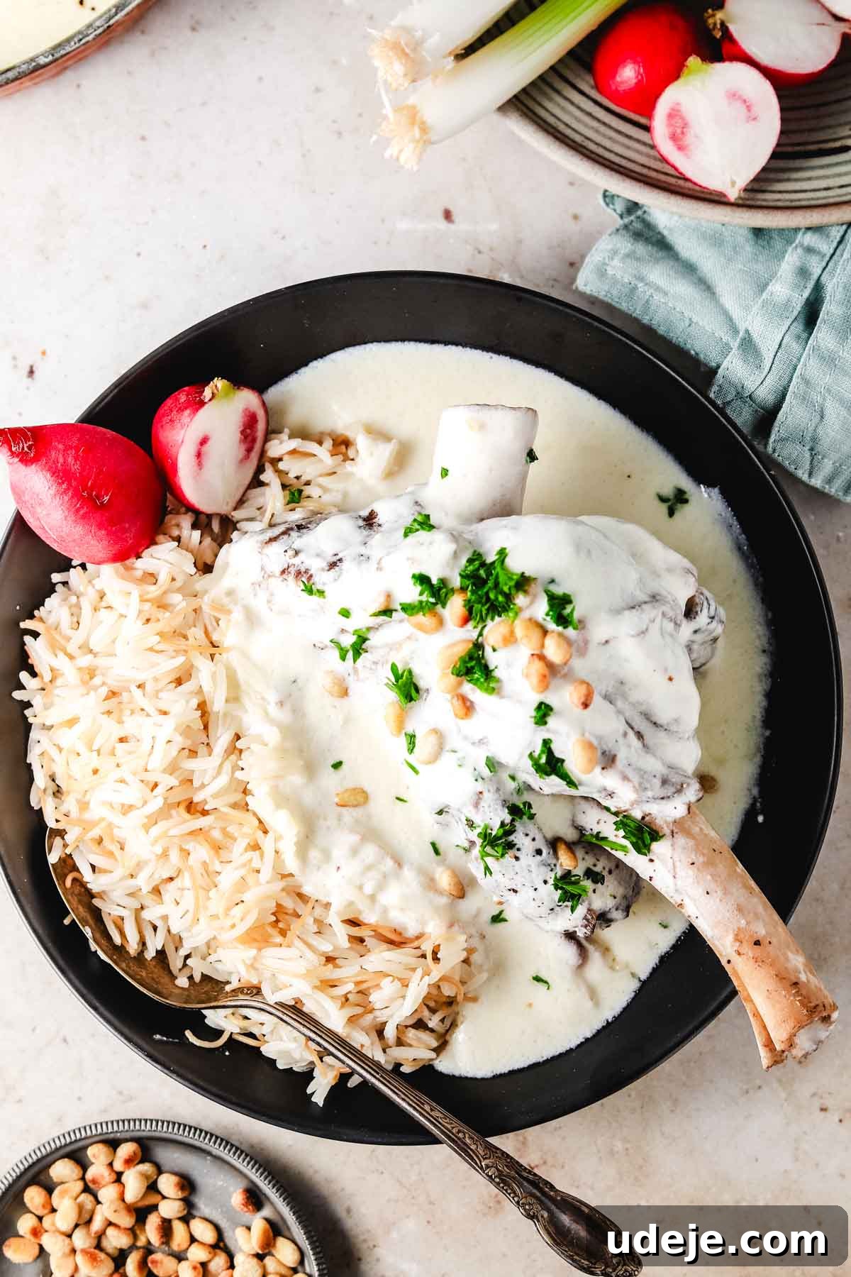 Syrian Shakriyeh: Velvety Lamb and Yogurt Stew 15 lamb shank in a yogurt sauce plated next to rice with vermicelli noodles in a black plate. The garnish is pine nuts, chopped parsley, and some cut up radish.