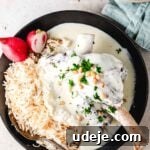Syrian Shakriyeh: Velvety Lamb and Yogurt Stew 25 lamb shank in a yogurt sauce plated next to rice with vermicelli noodles in a black plate. The garnish is pine nuts, chopped parsley, and some cut up radish.