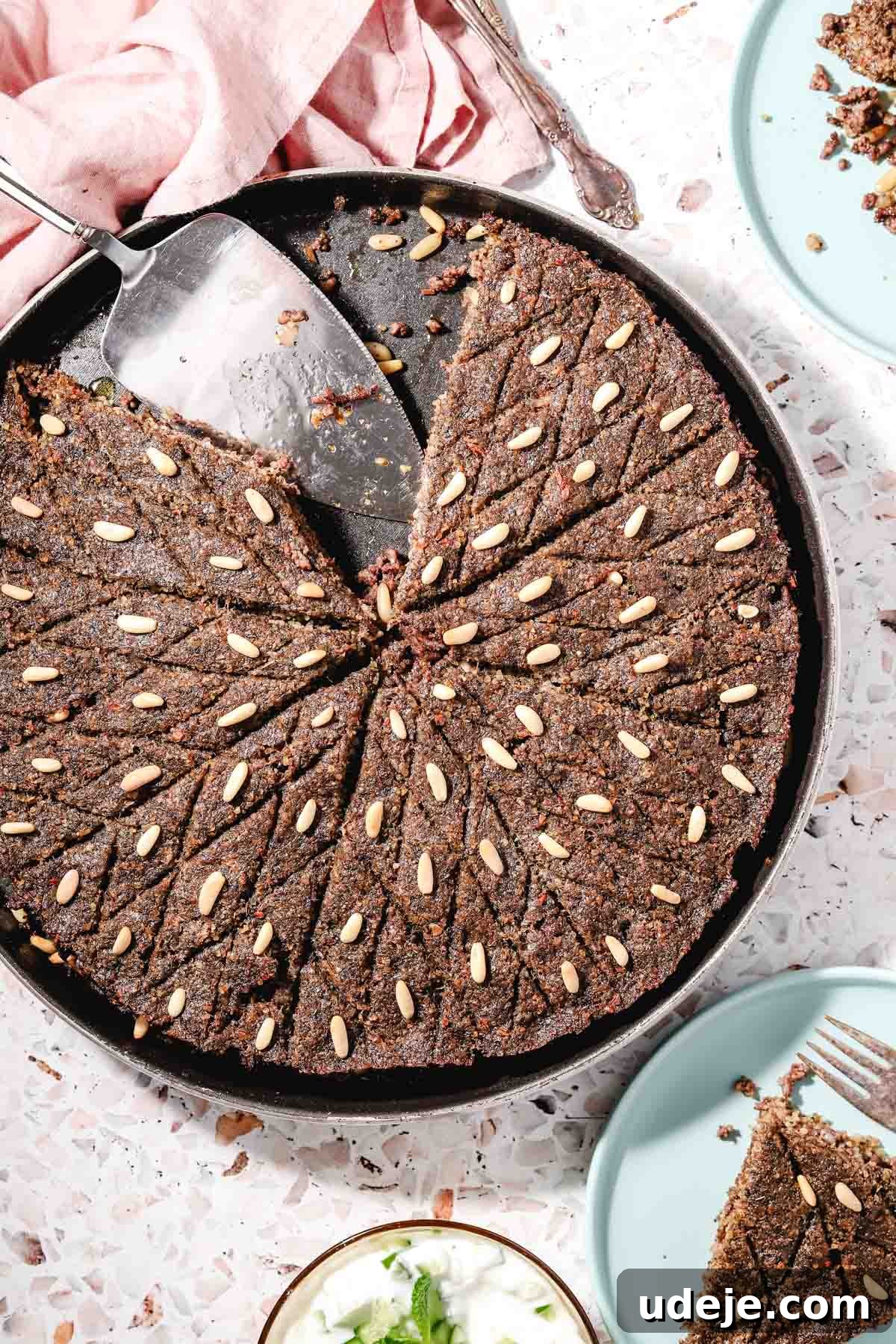 Baked Kibbeh Casserole 2 Golden-baked Kibbeh bil Sanieh in a large circular baking tray with a few slices already served, showcasing its flavorful layers. A metal cake server rests in the dish, set against a stylish pink terrazzo background with a soft pink towel in the corner. Adjacent, light blue plates with individual kibbeh pieces and cutlery, alongside a small bowl of refreshing yogurt garnished with mint leaves, complete the inviting scene.