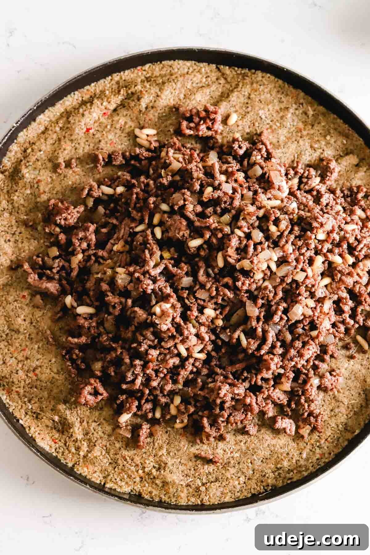 Baked Kibbeh Casserole 16 Sautéed ground beef and onions being placed meticulously on top of the kibbeh dough in a large circular baking tray on a light marble background.