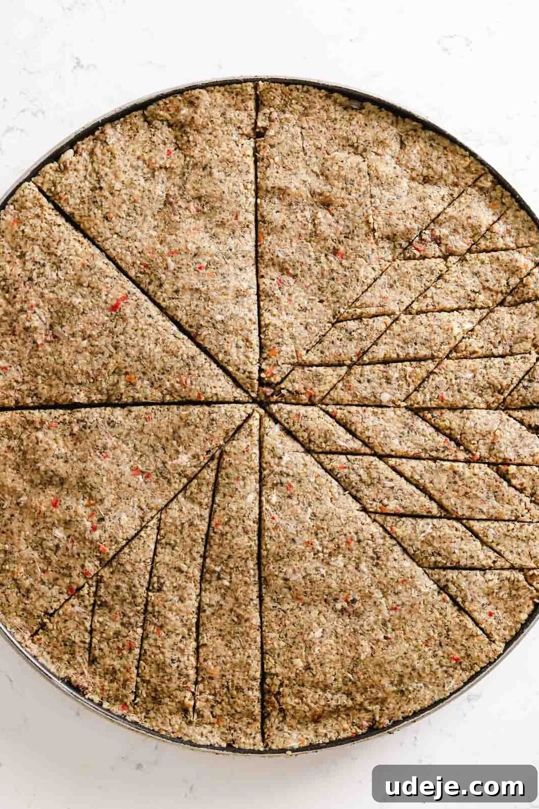 Baked Kibbeh Casserole 21 Unbaked Kibbeh bil Sanieh with intricate diamond shapes scored on half of its surface within the triangular sections in a large circular baking pan on a light marble surface.