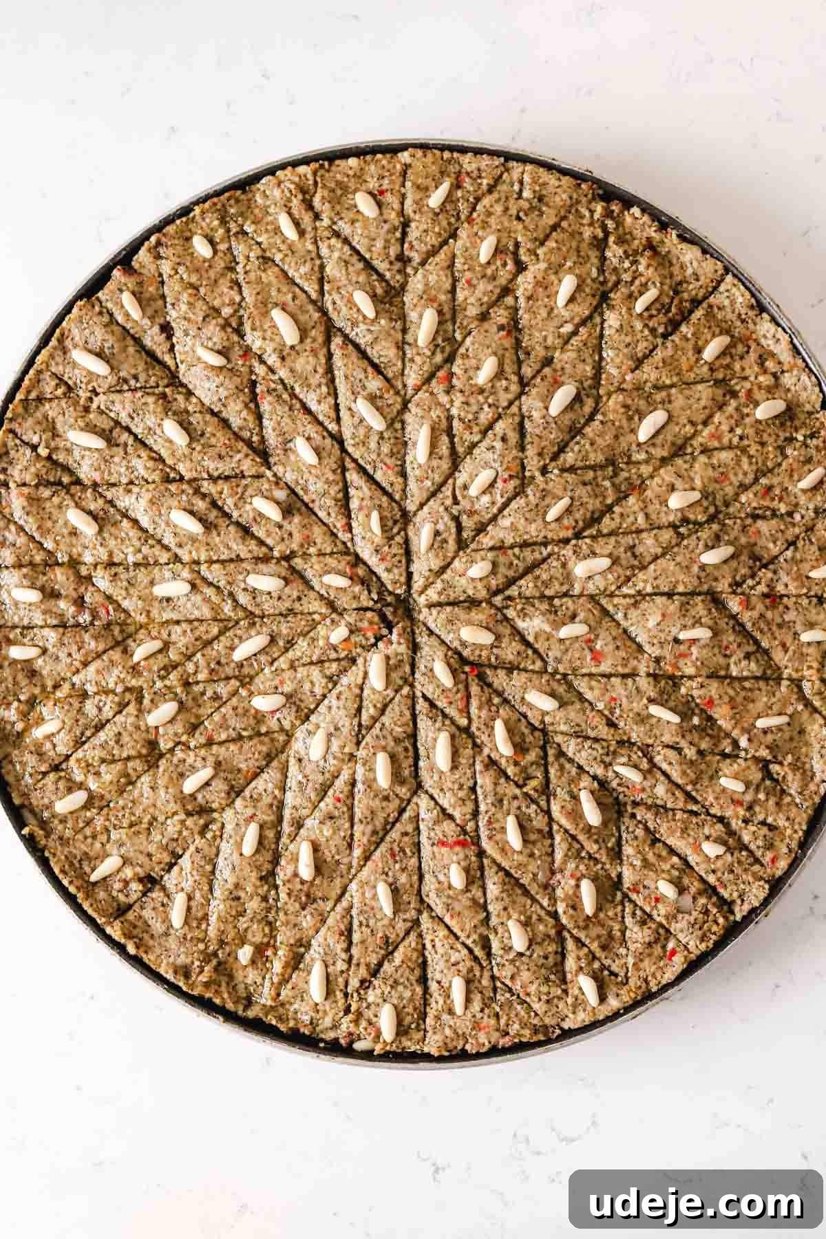 Baked Kibbeh Casserole 24 Unbaked Kibbeh bil Sanieh, neatly cut into diamond shapes, with a pine nut placed on each piece, set in a large circular baking pan on a light marble surface.