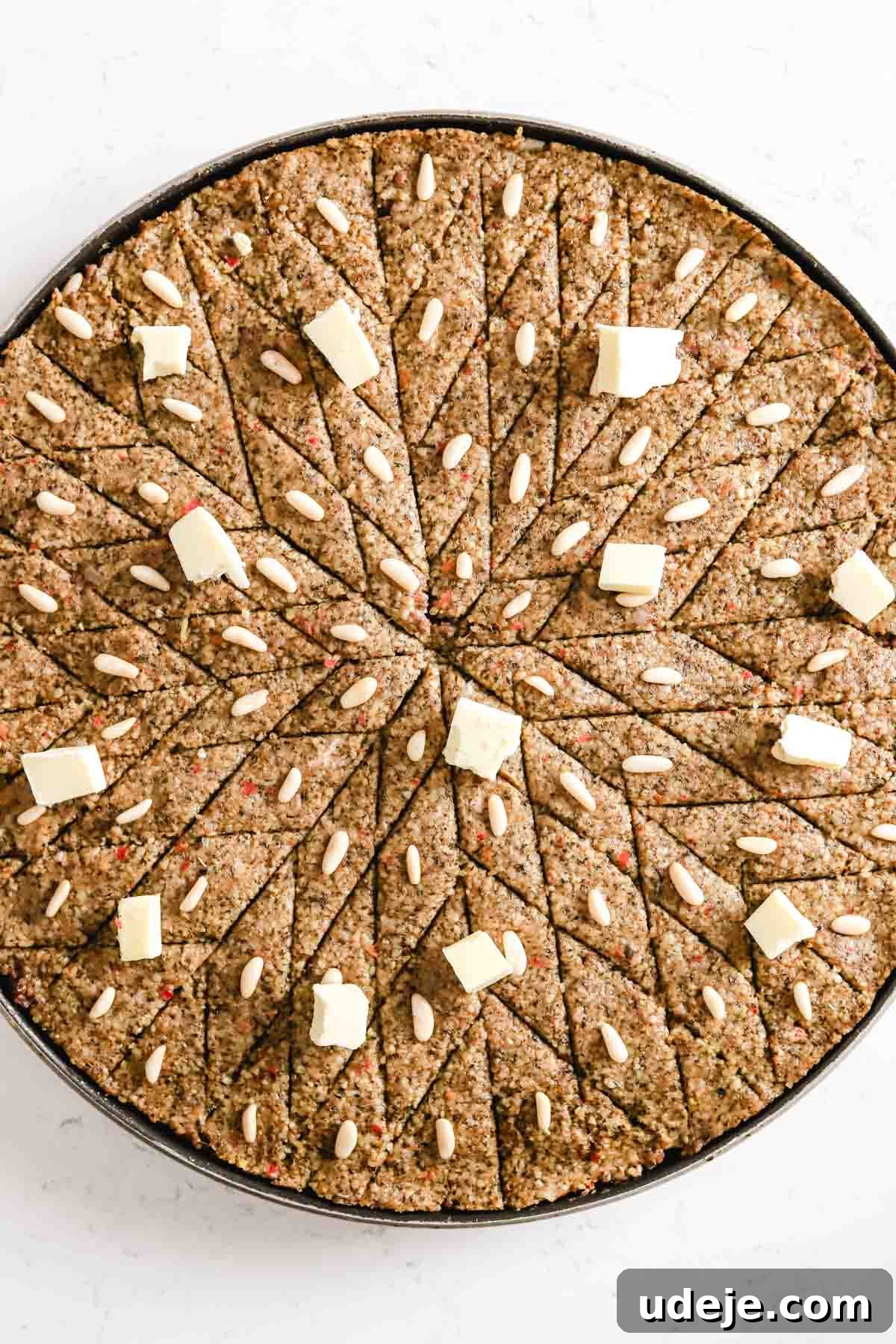 Baked Kibbeh Casserole 25 Unbaked Kibbeh bil Sanieh, scored into diamond shapes and adorned with pine nuts, receives a scattering of cold butter pieces in a large circular baking pan on a light marble surface, ready for baking.