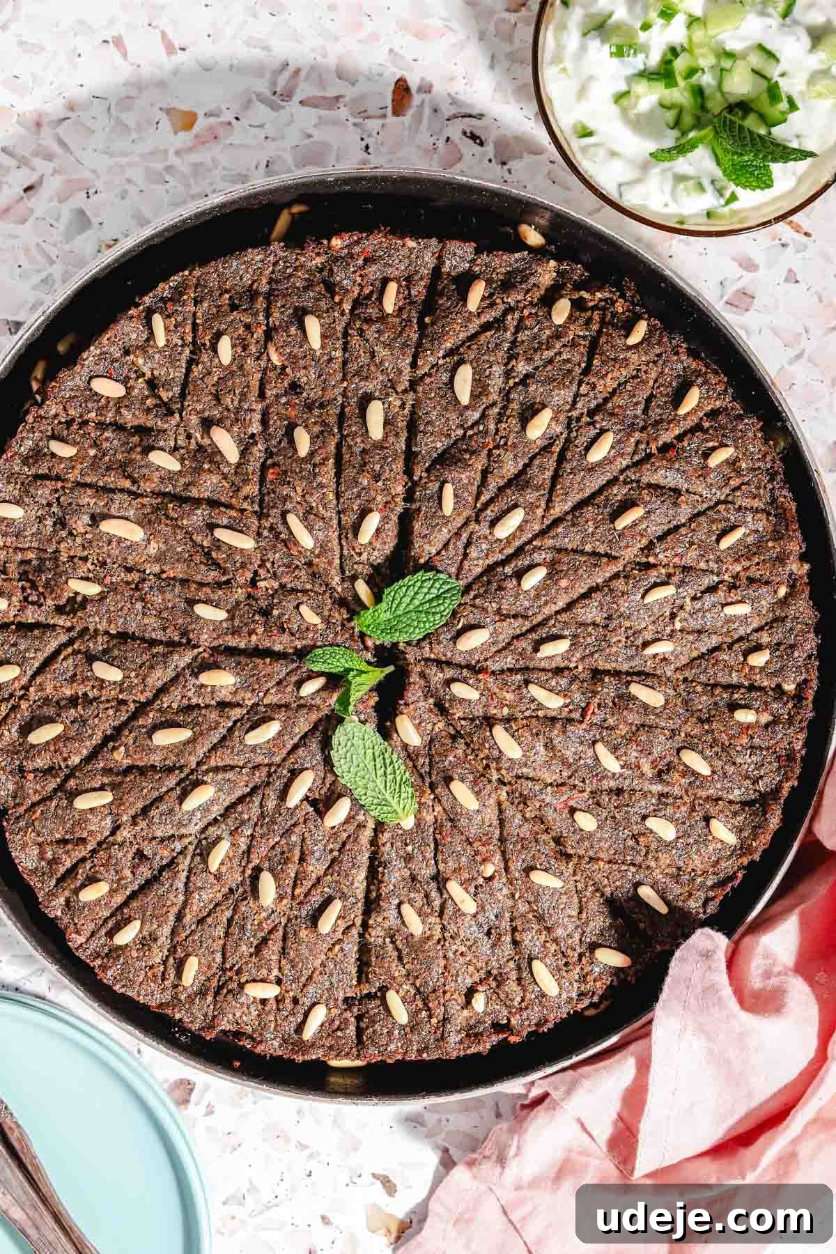 Baked Kibbeh Casserole 26 Perfectly baked Kibbeh bil Sanieh in a large circular baking tray, gleaming golden on a pink terrazzo background. A soft pink towel accents the corner, while light blue plates with cutlery and a small bowl of refreshing yogurt with mint leaves sit nearby, inviting a delightful meal.