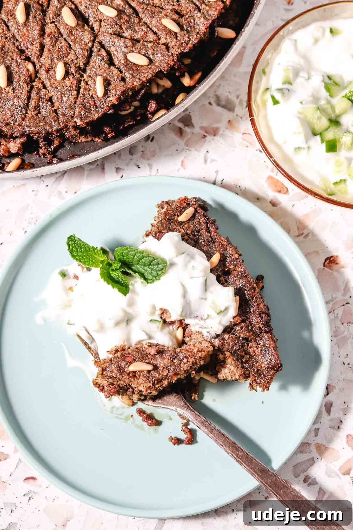 Baked Kibbeh Casserole 27 A slice of Kibbeh bil Sanieh gracefully plated on a light blue dish with a fork, elegantly topped with a dollop of yogurt and fresh mint. The dish is set on a pink terrazzo background, with a larger tray of kibbeh and a small bowl of refreshing cucumber yogurt salad in the background, completing the serving presentation.