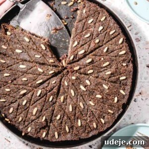 Golden-baked Kibbeh bil Sanieh in a large circular baking tray with a few slices already served, showcasing its flavorful layers. A metal cake server rests in the dish, set against a stylish pink terrazzo background with a soft pink towel in the corner. Adjacent, light blue plates with individual kibbeh pieces and cutlery, alongside a small bowl of refreshing yogurt garnished with mint leaves.