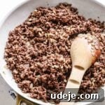 Sautéed ground beef with onions in a light grey skillet with gold handles with a wooden spoon on a blue and white towel on a light coloured background