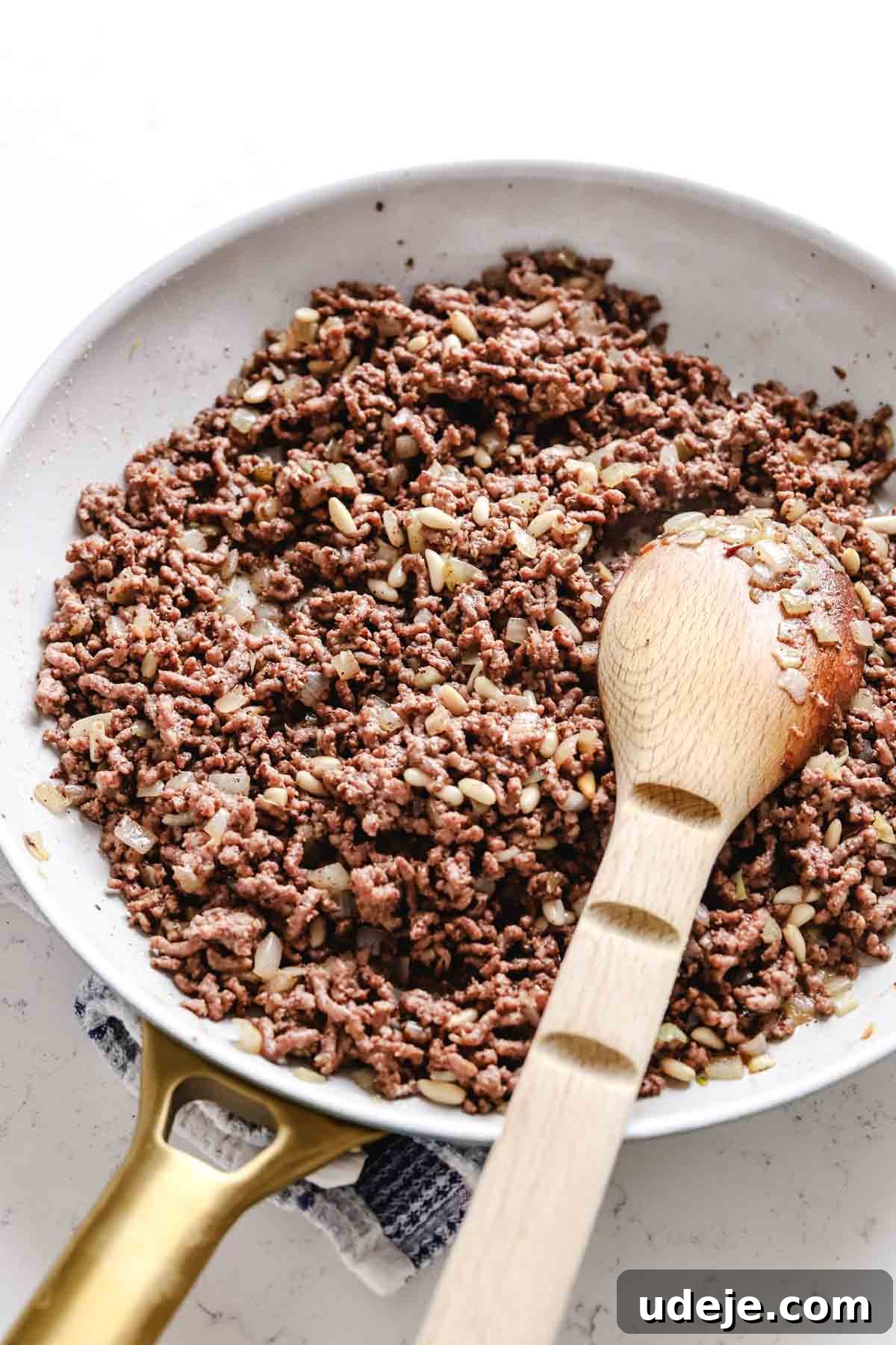 Baked Kibbeh Casserole 6 Sautéed ground beef with onions simmering in a light grey skillet with elegant gold handles. A wooden spoon rests in the pan, set on a blue and white towel against a light-colored background, illustrating the preparation of the savory hashweh filling.