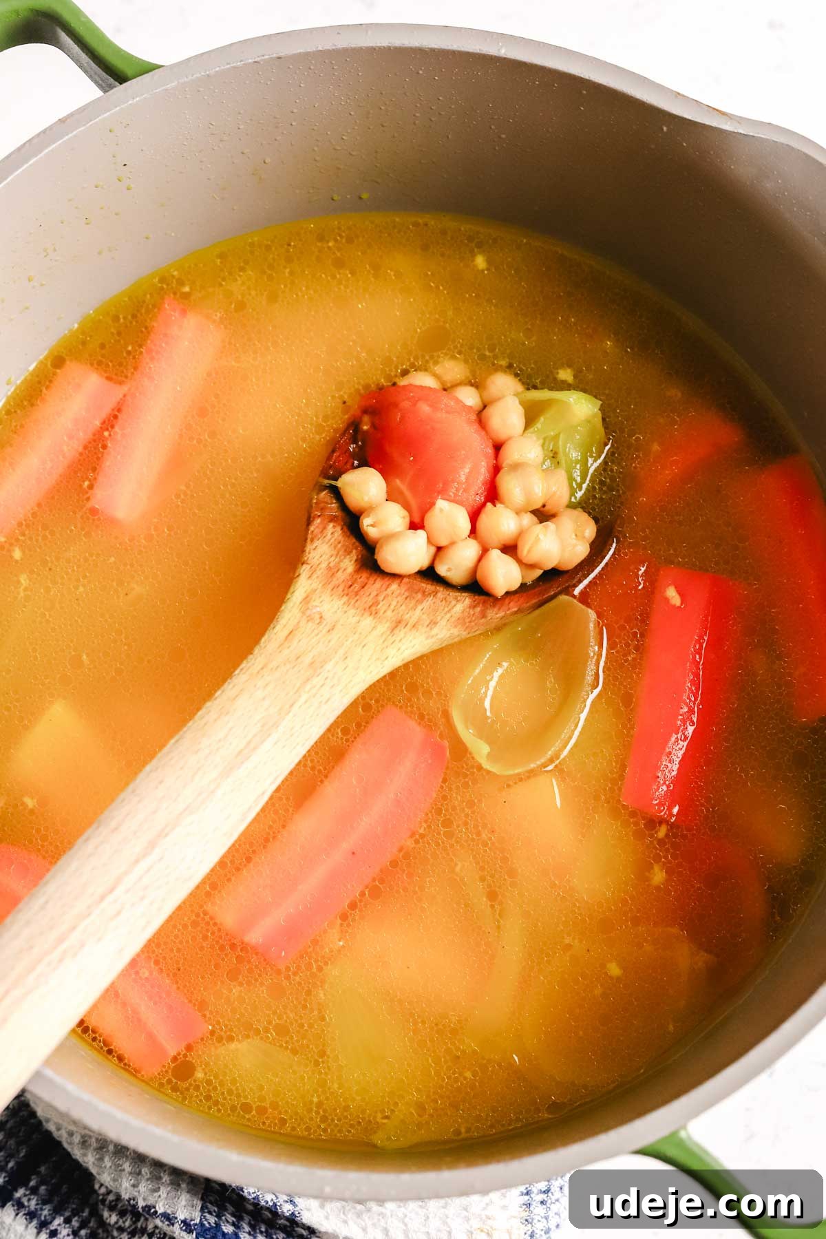 A large pot holds a simmering chicken broth generously filled with colorful vegetables. A wooden spoon rests inside, scooping up chickpeas and a medley of vegetables, showcasing the hearty contents of the stew.