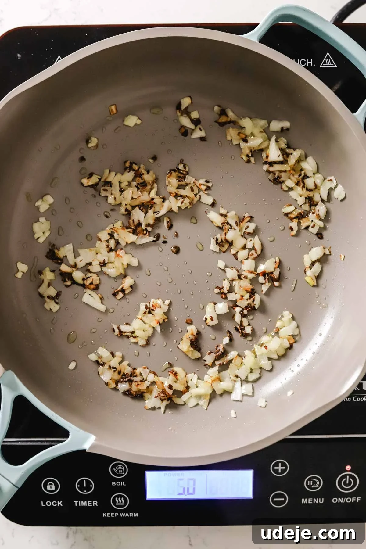Finely diced onions and fragrant dill seeds are being gently softened in a nonstick skillet, creating an aromatic base for the maftoul.