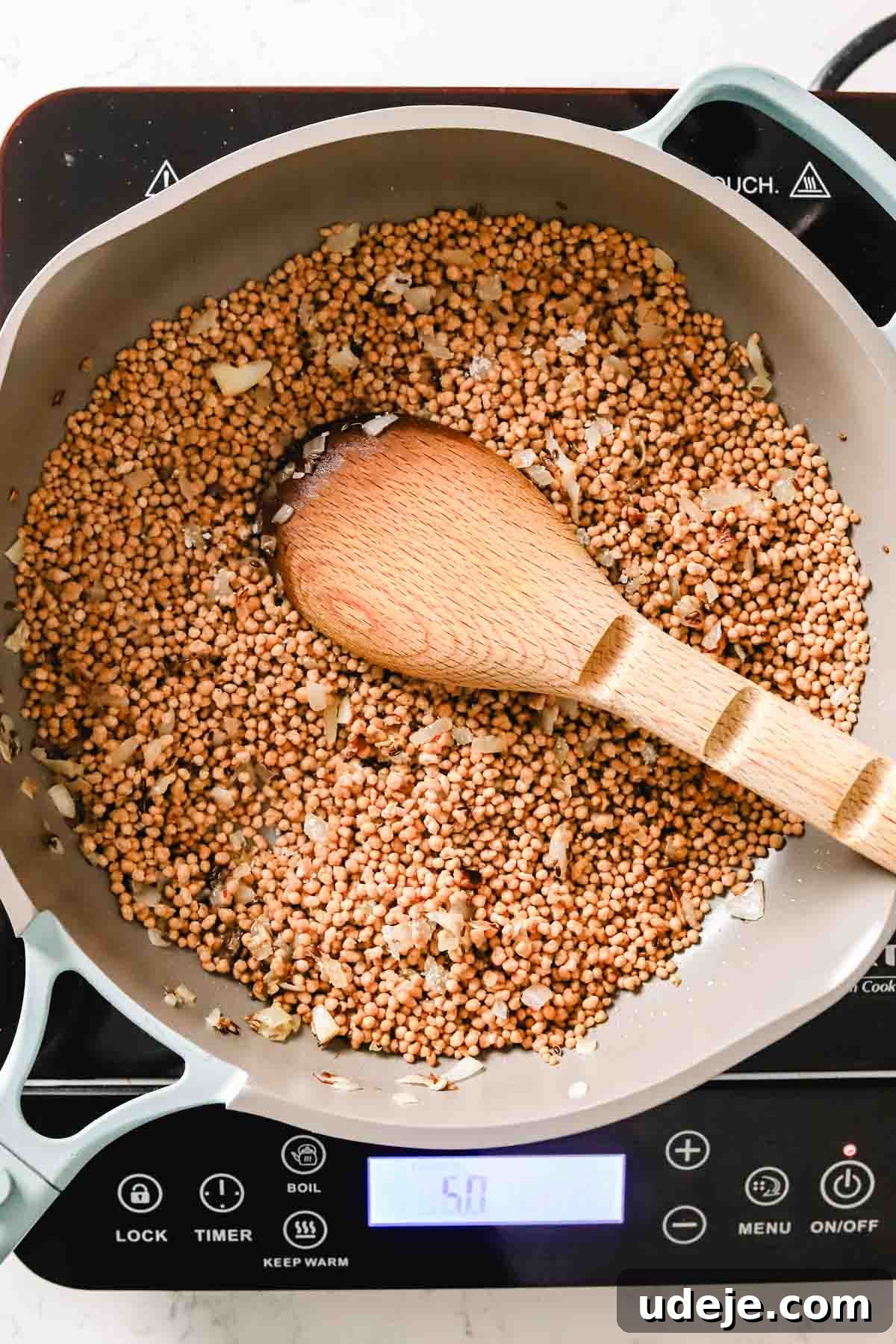 Palestinian maftoul, along with finely diced onions and fragrant dill seeds, is being stirred in a nonstick skillet with a wooden spoon, preparing the grains for cooking.