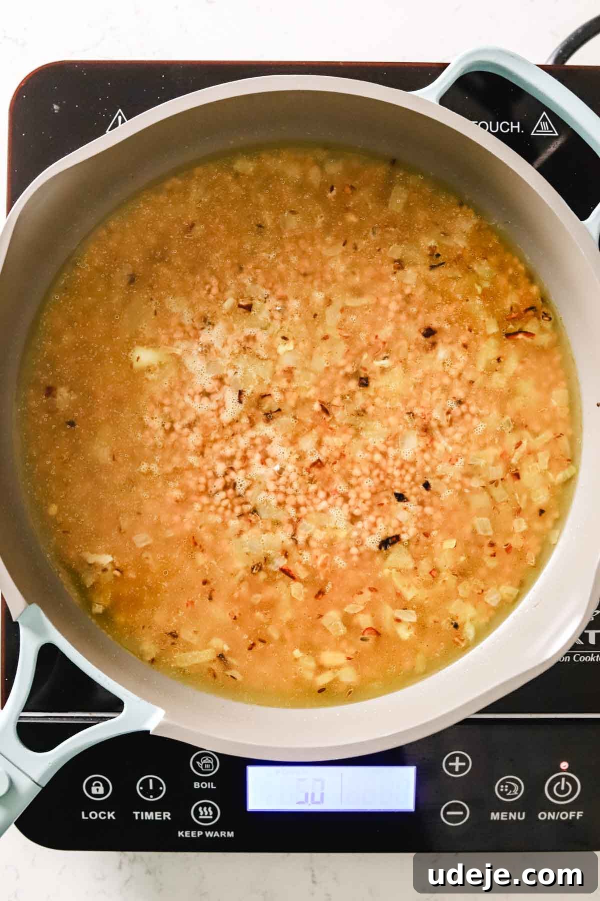 Palestinian maftoul simmering in a nonstick skillet with diced onions, dill seeds, and chicken broth, absorbing the liquid to achieve perfect tenderness.