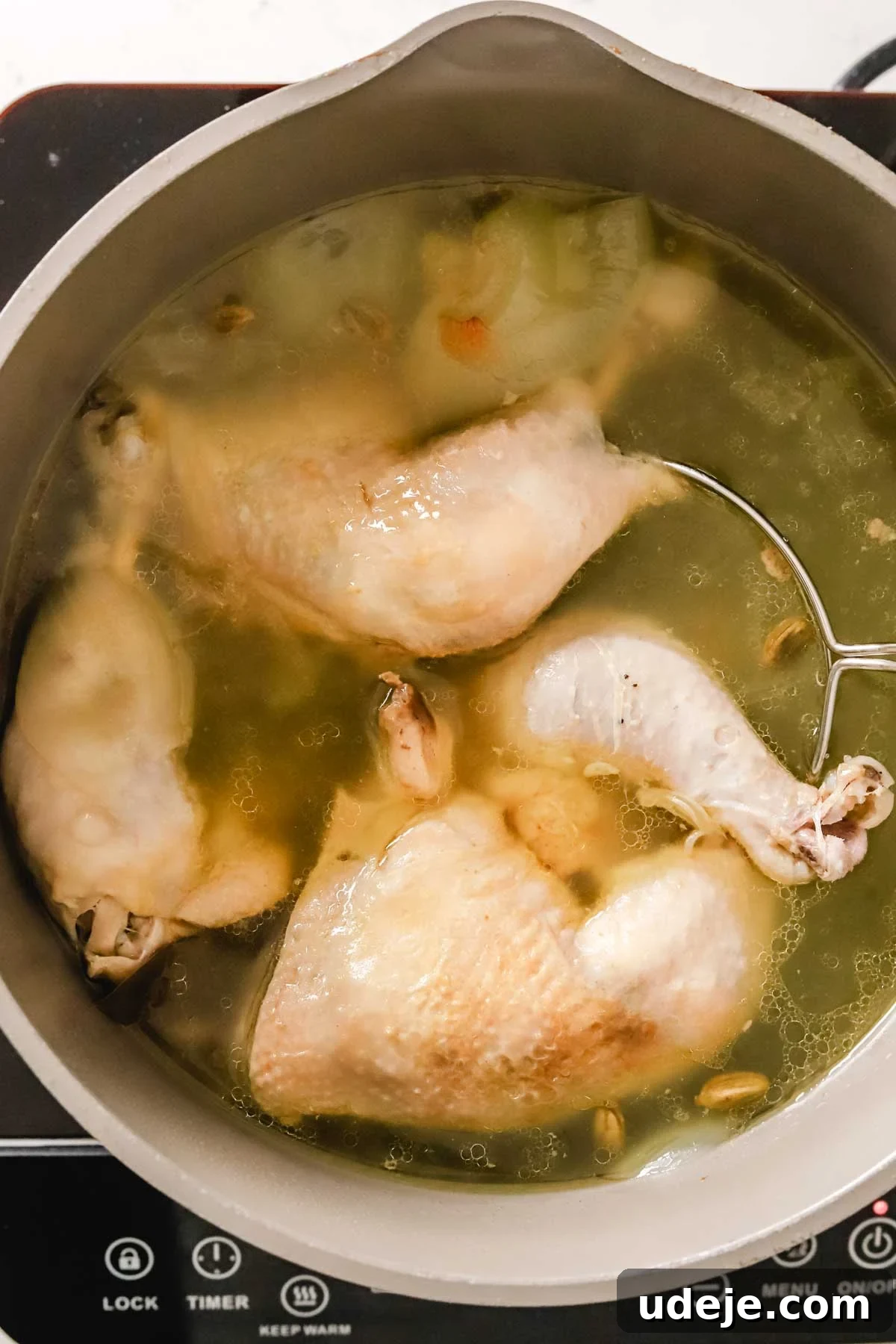 Cooked bone-in chicken legs and flavorful broth simmer quietly in a pot, accompanied by whole spices and quartered onions, forming the aromatic base for the maftoul dish.