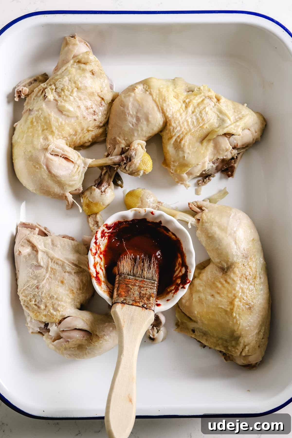 Boiled bone-in chicken legs are arranged on a white oven tray, with a small bowl containing tomato paste and a pastry brush nearby, indicating preparation for broiling.