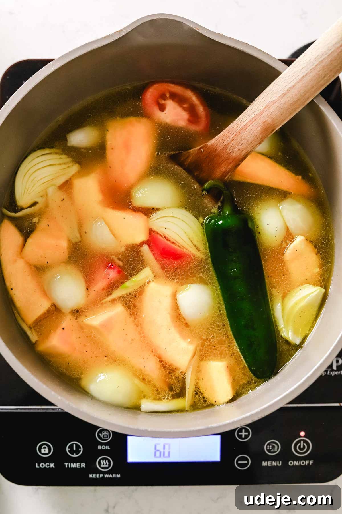 A rich chicken broth simmers in a pot, brimming with large, vibrant chunks of carrots, onions, squash, and tomato, along with a single large jalapeno, indicating a flavorful stew in progress.