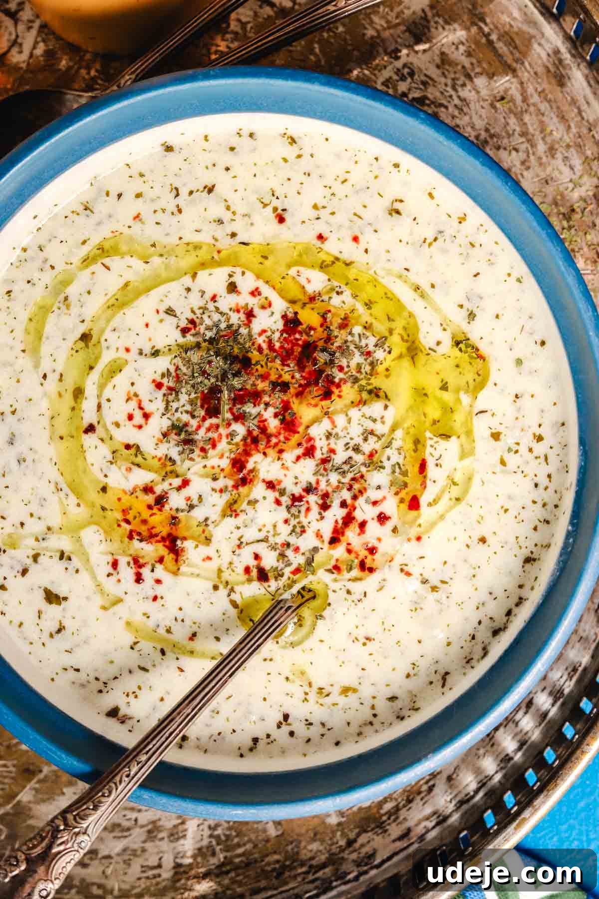 A vibrant blue bowl filled with creamy Yayla Corbasi, a Turkish yogurt soup, garnished with dried mint, a sprinkle of red Aleppo pepper, and a glistening drizzle of olive oil, with a soup spoon resting in the bowl.