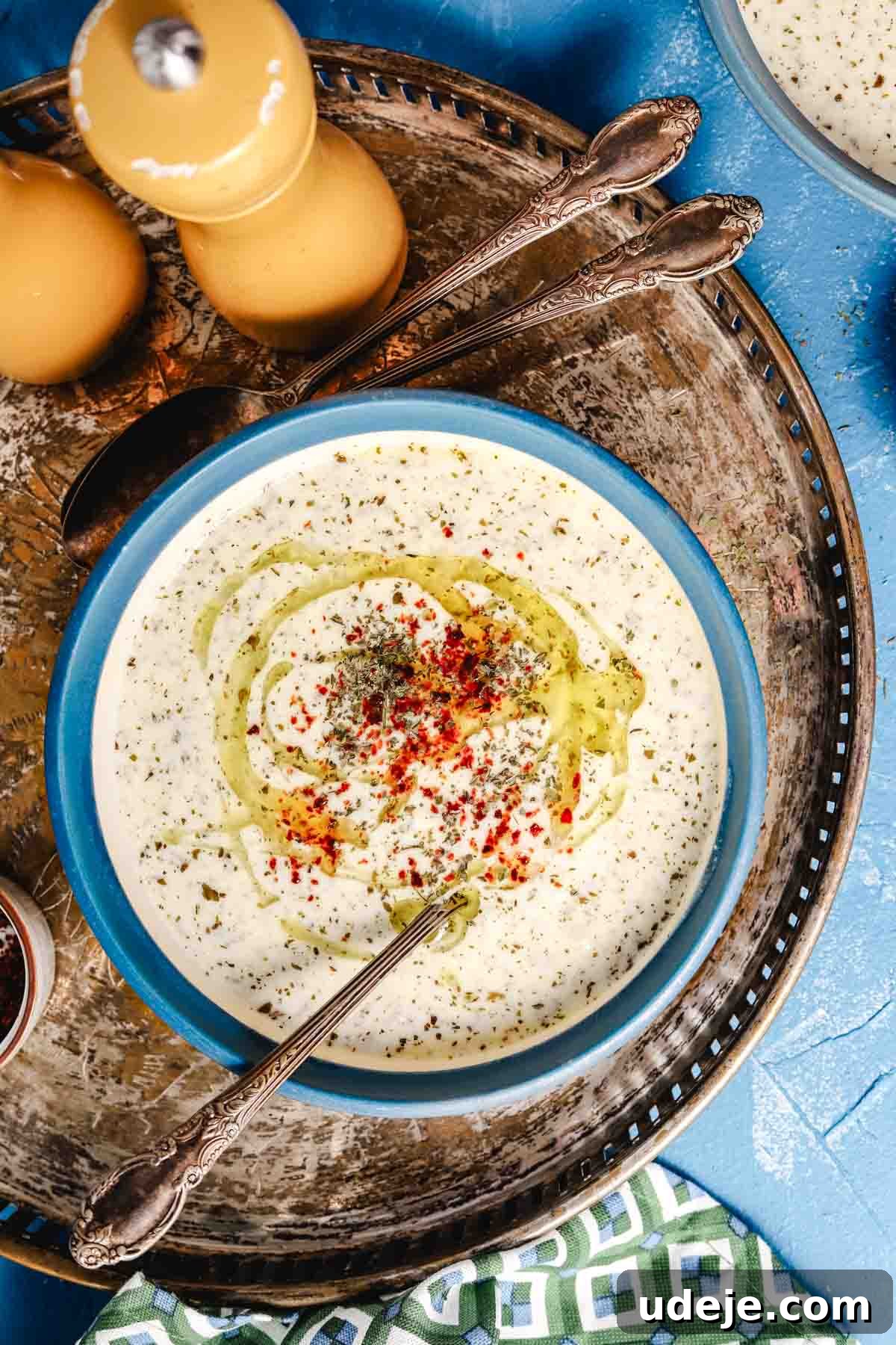 Yayla Corbasi, a traditional Turkish yogurt soup, beautifully presented in a blue ceramic bowl on a large metal serving tray, flanked by yellow salt and pepper mills and elegant soup spoons.