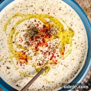 yayla corbasi which is yogurt soup is served in a blue bowl. The soup is garnished with dried mint, aleppo pepper and olive oil, with a soup spoon inside the bowl