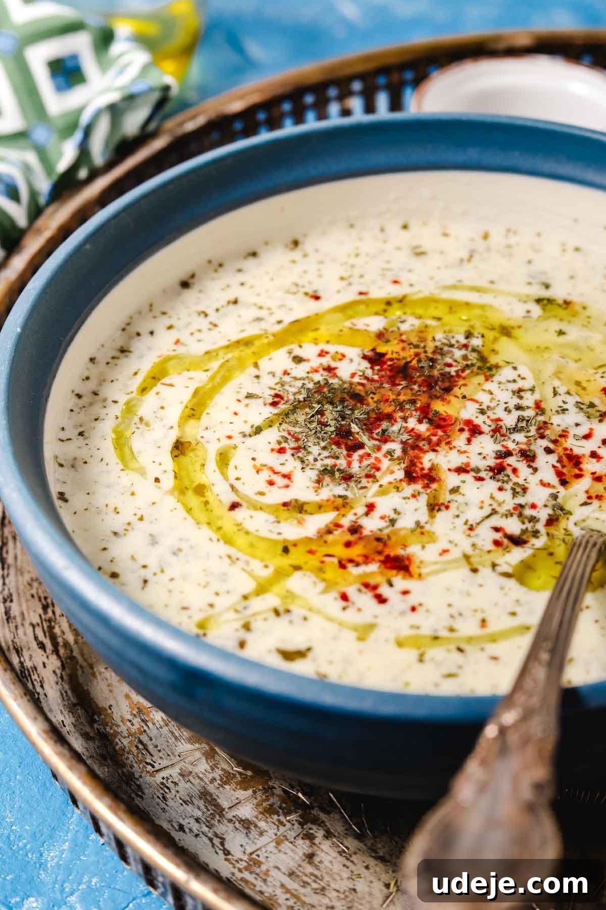 A close-up shot of Yayla Corbasi, a Turkish yogurt soup, elegantly served in a blue ceramic bowl. The soup is beautifully garnished with vibrant dried mint, a touch of spicy Aleppo pepper, and a delicate swirl of olive oil, with a silver soup spoon invitingly placed inside the bowl.