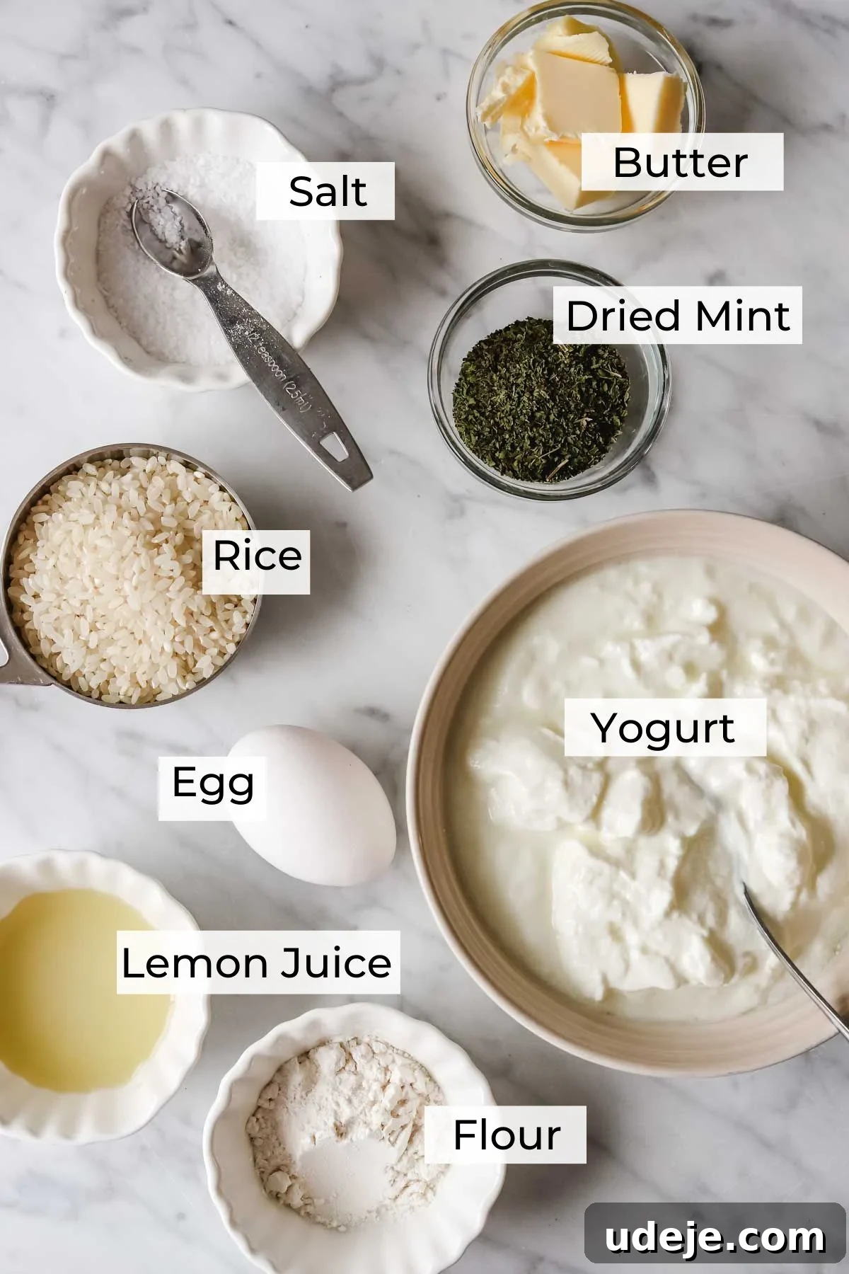 A flat lay photograph showcasing the simple yet essential ingredients for Yayla Corbasi: creamy yogurt, uncooked white rice, a fresh egg, vibrant lemon juice, aromatic dried mint, golden butter, and a pinch of salt, artfully arranged on a light background.