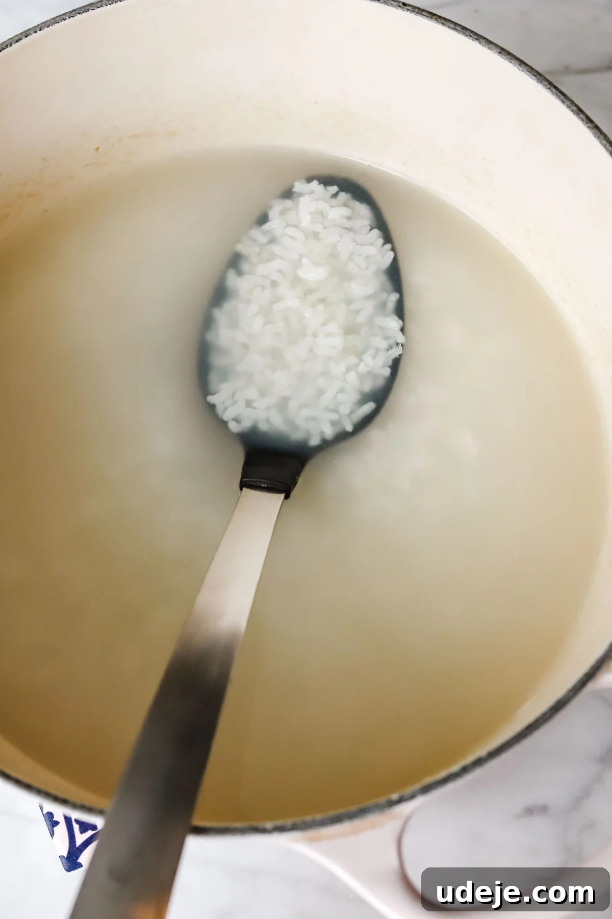 A close-up shot of a soup pot filled with cooked white rice floating in water, with a large serving spoon dipped into the tender grains.
