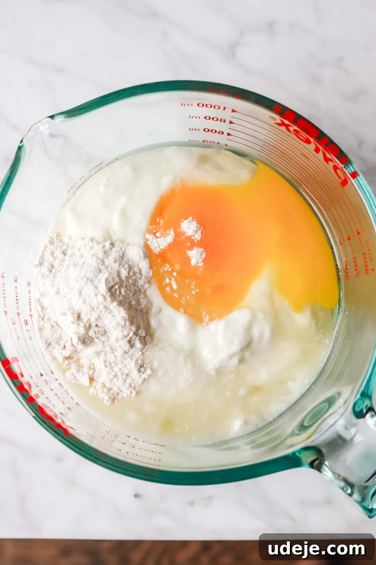 A clear measuring cup showcasing the essential ingredients for the yogurt base of Yayla Corbasi: creamy yogurt, a raw cracked egg, a scoop of cornstarch (or flour), and a splash of fresh lemon juice.