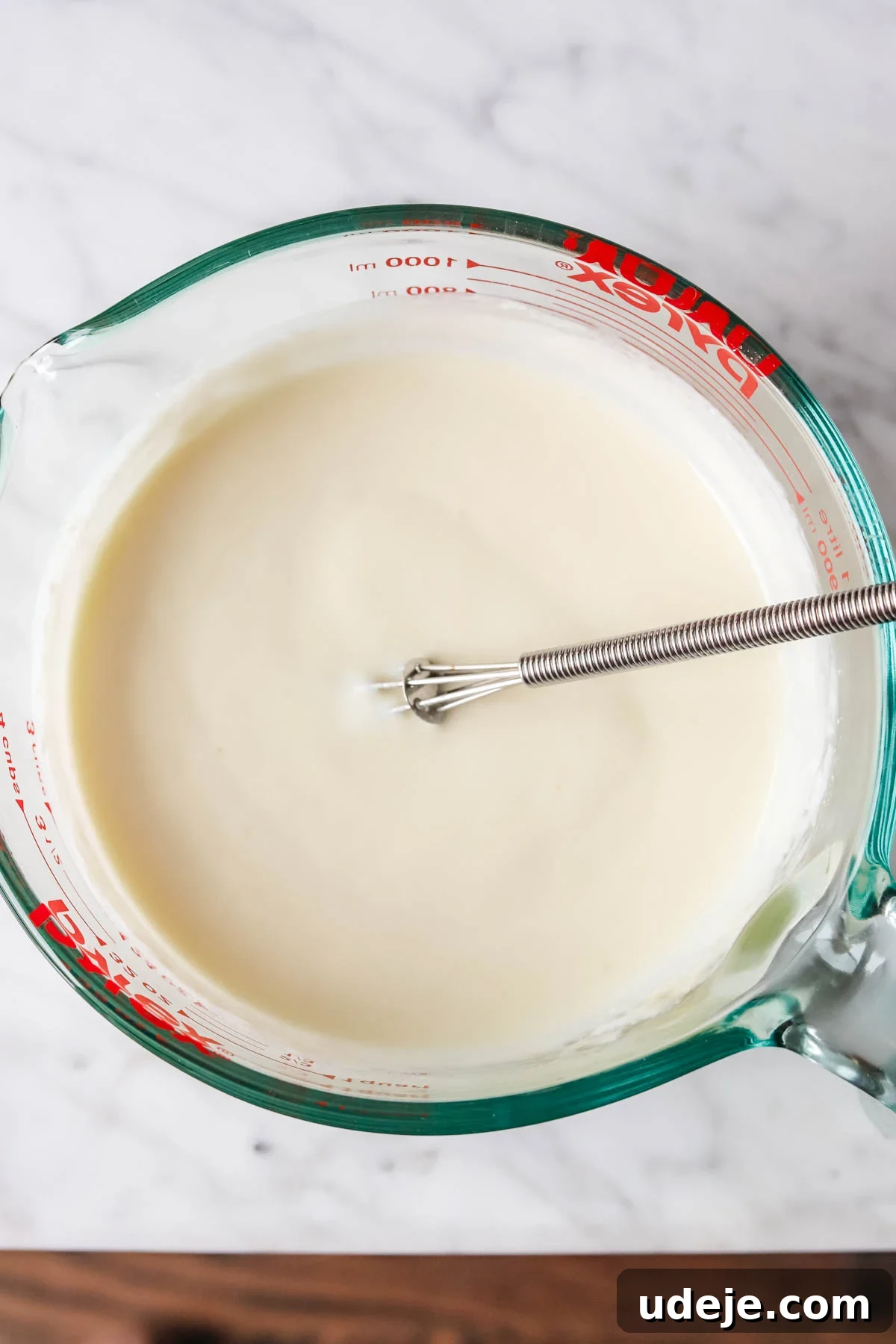 A measuring cup containing a thick, smooth white liquid, a mixture of whisked yogurt, a raw egg, flour, and lemon juice, with a small whisk immersed.