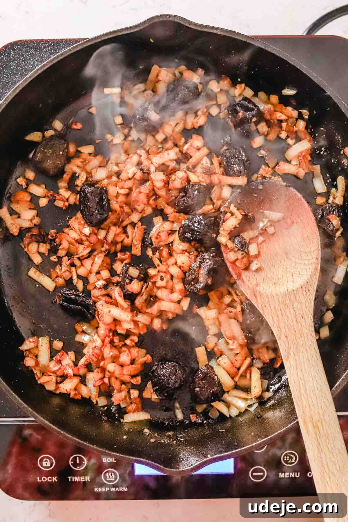 Golden Crispy Moroccan Oven Chicken 11 chopped onions being sautéed in a large cast iron pan with prunes on a hot plate with a wooden spoon on a white marble background