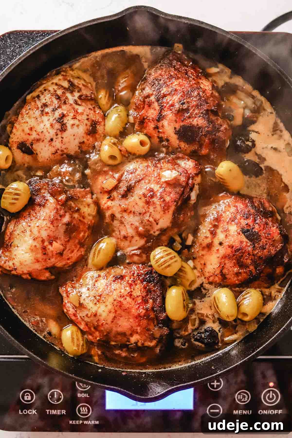 Golden Crispy Moroccan Oven Chicken 13 Seared chicken in broth with prunes and olives in a large cast iron pan on a hot plate on a white marble background