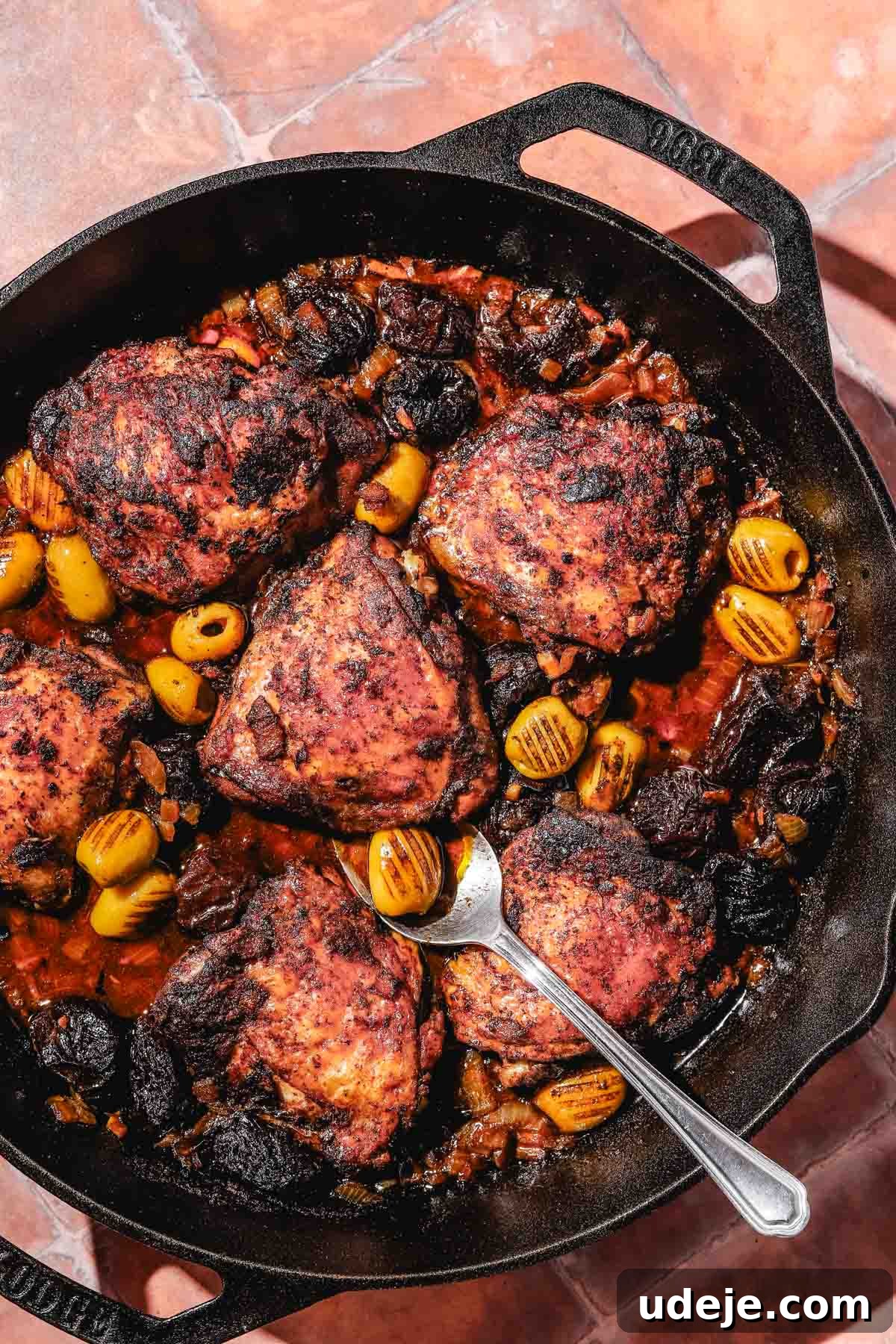 Golden Crispy Moroccan Oven Chicken 14 Moroccan Chicken in a large cast iron pan with a metal spoon on a rust tiled background