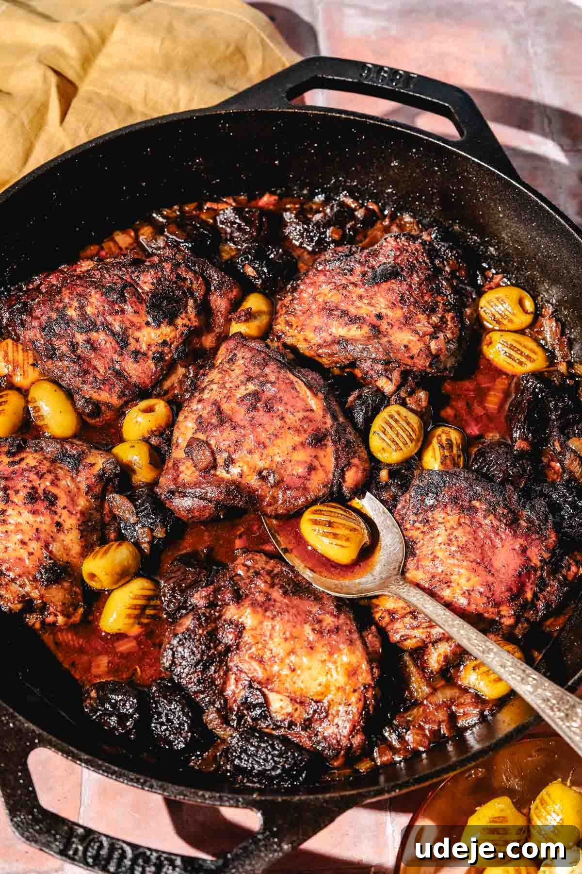 Golden Crispy Moroccan Oven Chicken 15 Moroccan Chicken in a large cast iron pan with a metal spoon, a yellow towel and a glass bowl with olives, on a rust tiled background