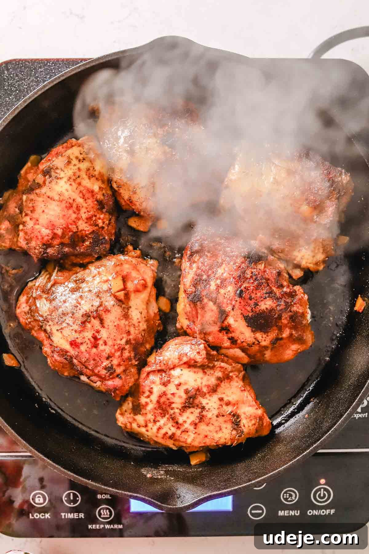 Golden Crispy Moroccan Oven Chicken 8 chicken thighs being seared in a cast iron pan on a hot plate on a white marble background
