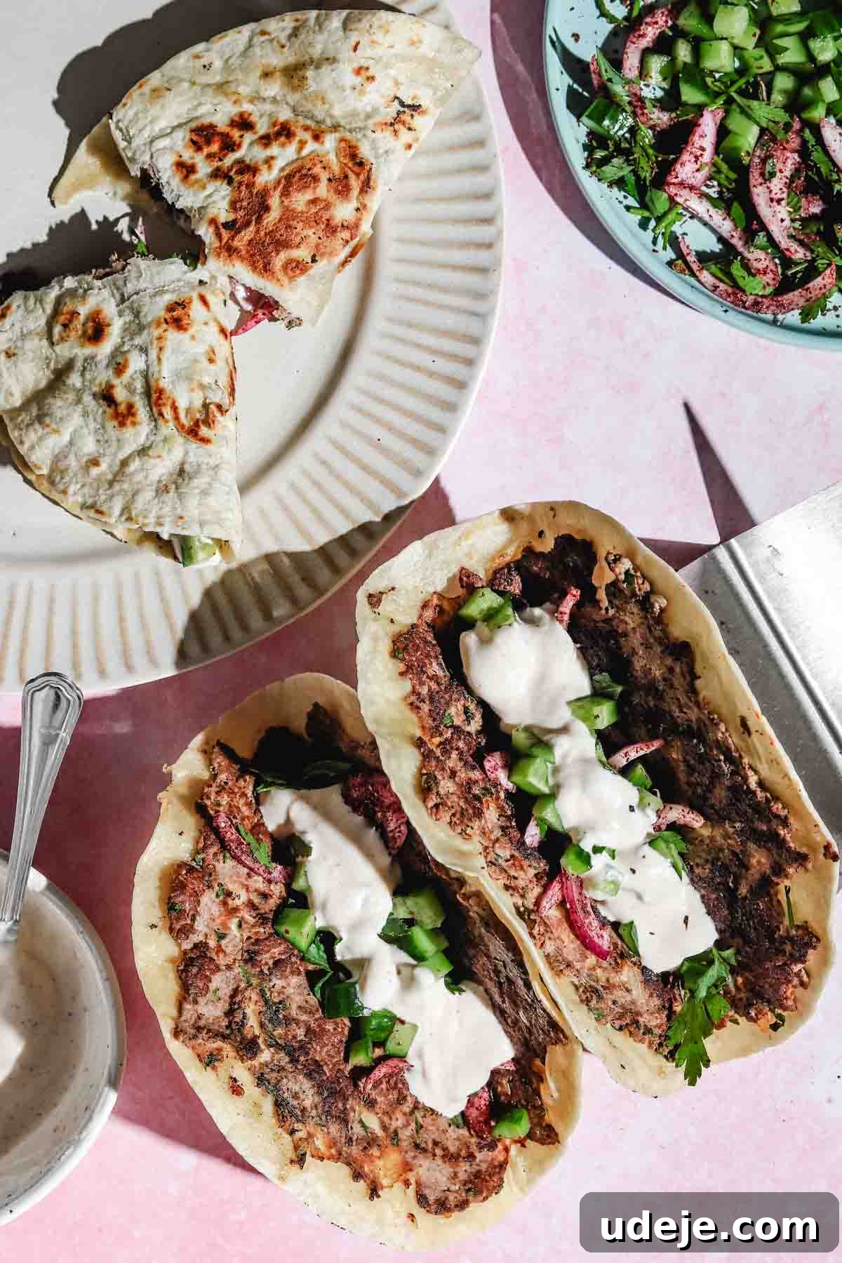 Two folded upright Kofta Smash Tacos topped with fresh cucumbers, red onions, and a creamy white sauce. A broken smash taco sits on a beige plate next to a vibrant cucumber onion salad in a blue bowl, and a small white bowl holding more white sauce with a metal spoon.