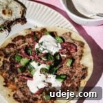 Kofta smash taco topped with cucumber and onions with white sauce on a beige plate with a small white bowl with white sauce on a pink background with a pink tea towel