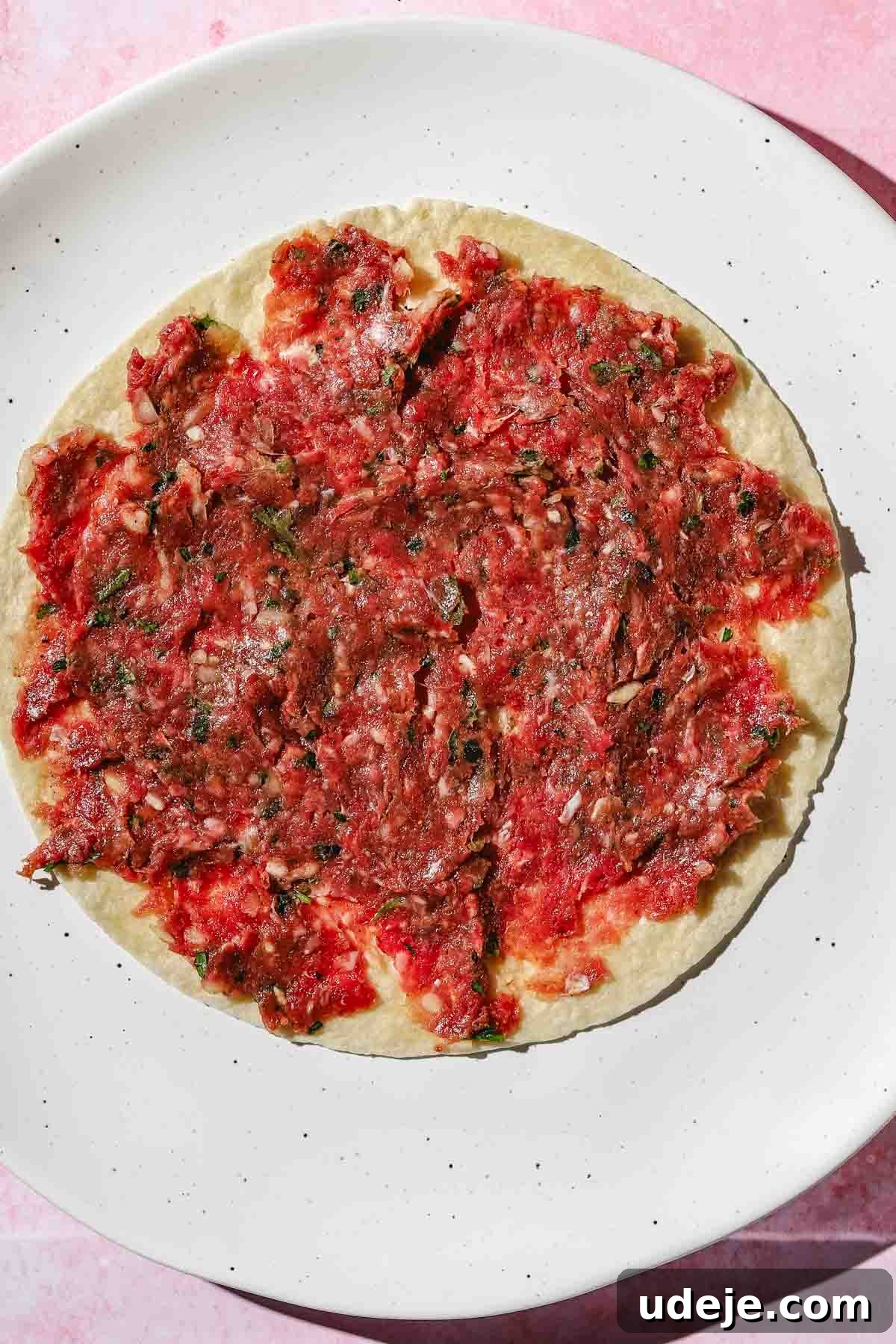 Ground beef kofta mixture spread thinly on a small tortilla, resting on a white speckled plate against a pink background.