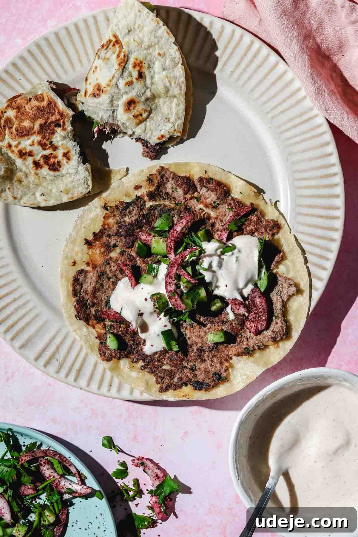 A Kofta Smash Taco topped with fresh cucumber, red onions, and a drizzle of white sauce, presented on a beige plate. Alongside are a small white bowl of white sauce and a blue bowl containing a red onion and parsley salad, all on a pink background with a matching tea towel.
