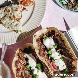 2 folded upright kofta smash tacos topped with cucumbers, red onions and white sauce with a folded smash taco broken in half on a beige plate and a cucumber onion salad in a blue bowl and a small white bowl with white sauce with a metal spoon